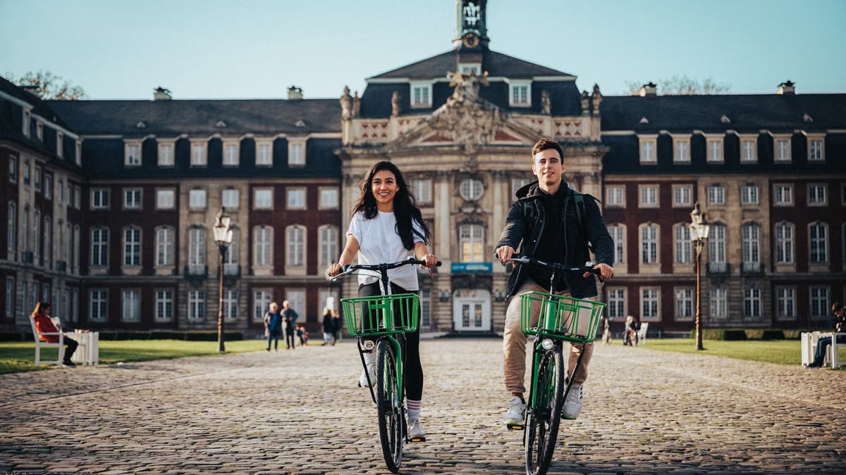 A couple rides bicycles in front of the University of Münster on a sunny day.